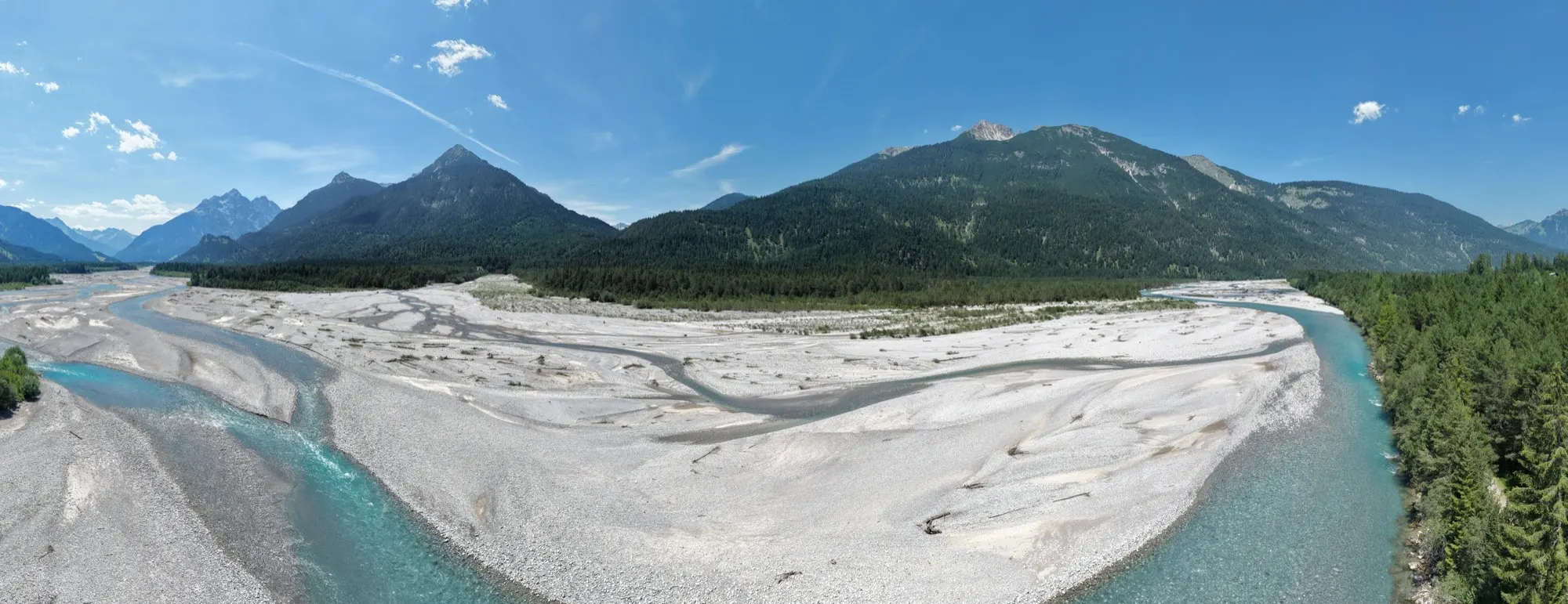 360-Grad-Panorama eines türkisfarbenen Gebirgsflusses in breitem Kiesbett zwischen bewaldeten Bergen