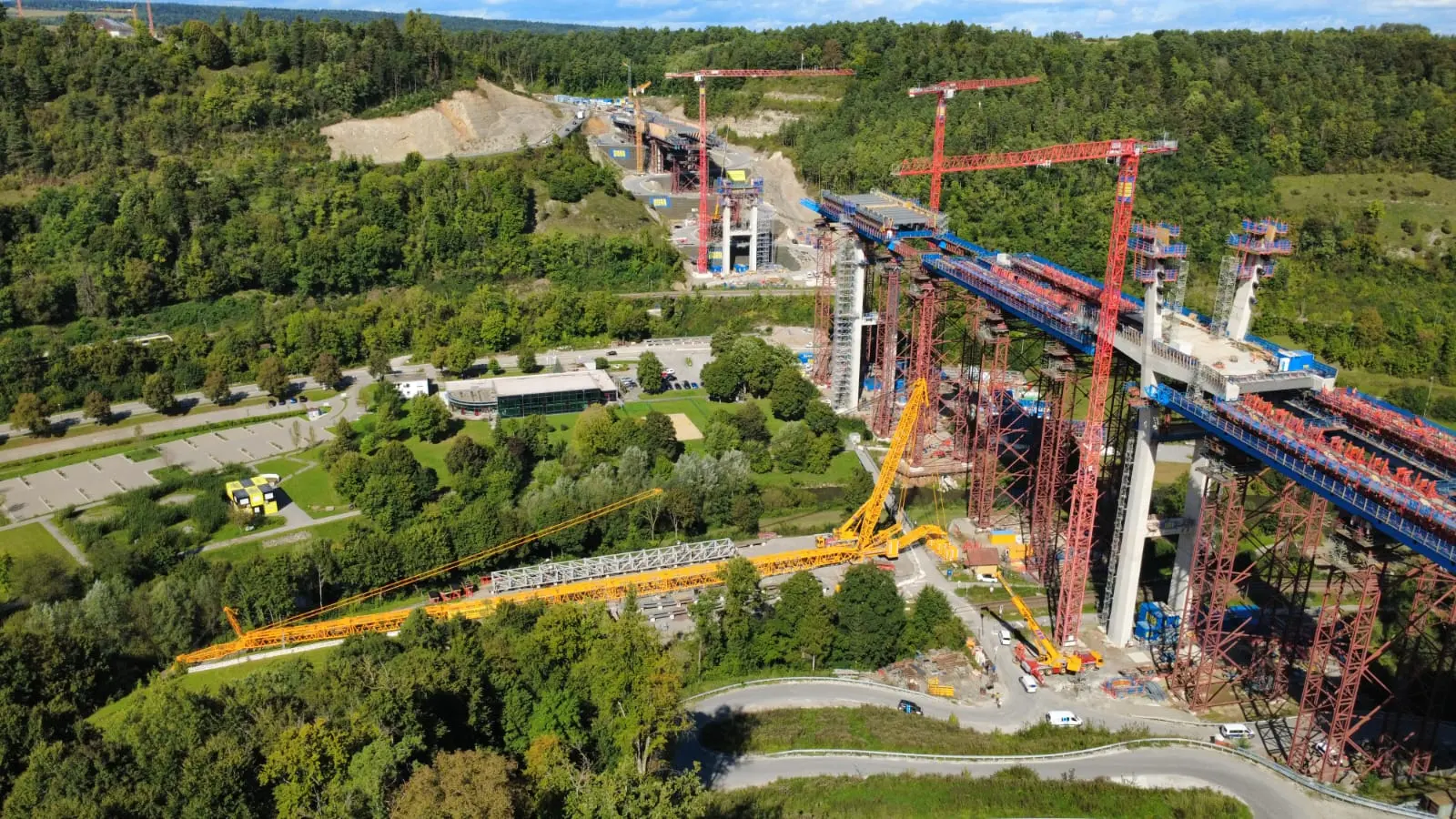Luftaufnahme einer großen Autobahnbrücke im Bau mit mehreren Baukränen über einem bewaldeten Tal
