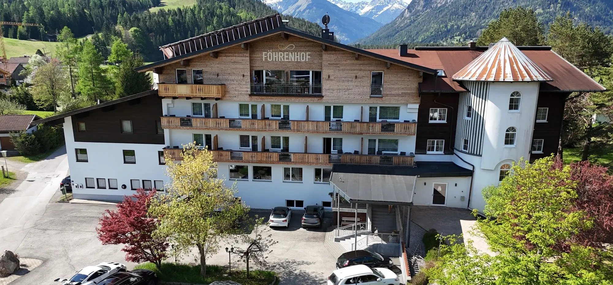 Luftaufnahme eines großen Hotelgebäudes im Alpenstil mit umliegenden Wiesen und Bergen